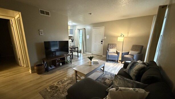 A living room with a sofa, coffee table, and TV. There is a door leading to another room. - Stay Longer for Less – Affordable Extended Stay (Garland)