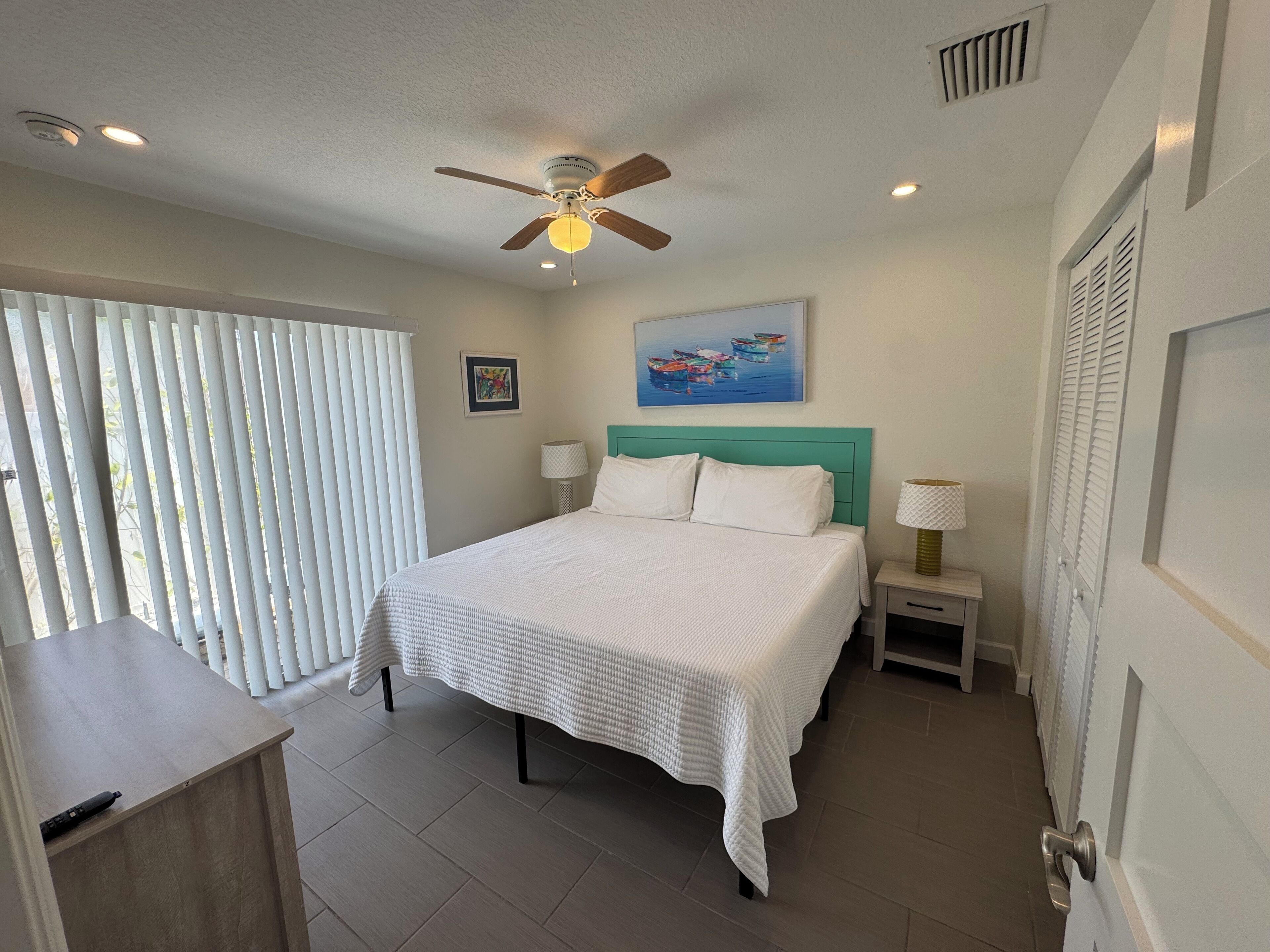 A bedroom with a bed, a ceiling fan, a painting, and a window with blinds.