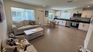 A modern living room with a sofa, coffee table, and dining area. The kitchen features white cabinets, a microwave, and a stainless steel oven.