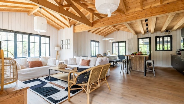 Amplia sala de estar con sofá, sillas y una mesa de centro. Cuenta con una cocina con barra y taburetes. La habitación tiene vigas de madera en el techo y ventanas grandes.