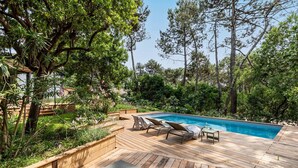 Una terraza de madera con piscina, rodeada de vegetación exuberante y altos árboles.