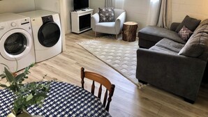 Kompakti olohuone, jossa on pyykinpesukone, harmaa sohva, pieni pöytä ruutukuosisella pöytäliinalla ja valkoisella jalustalla varustettu televisio.