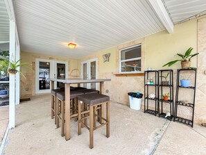Un patio cubierto con mesa y sillas, un cubo de basura y un estante con plantas en macetas.