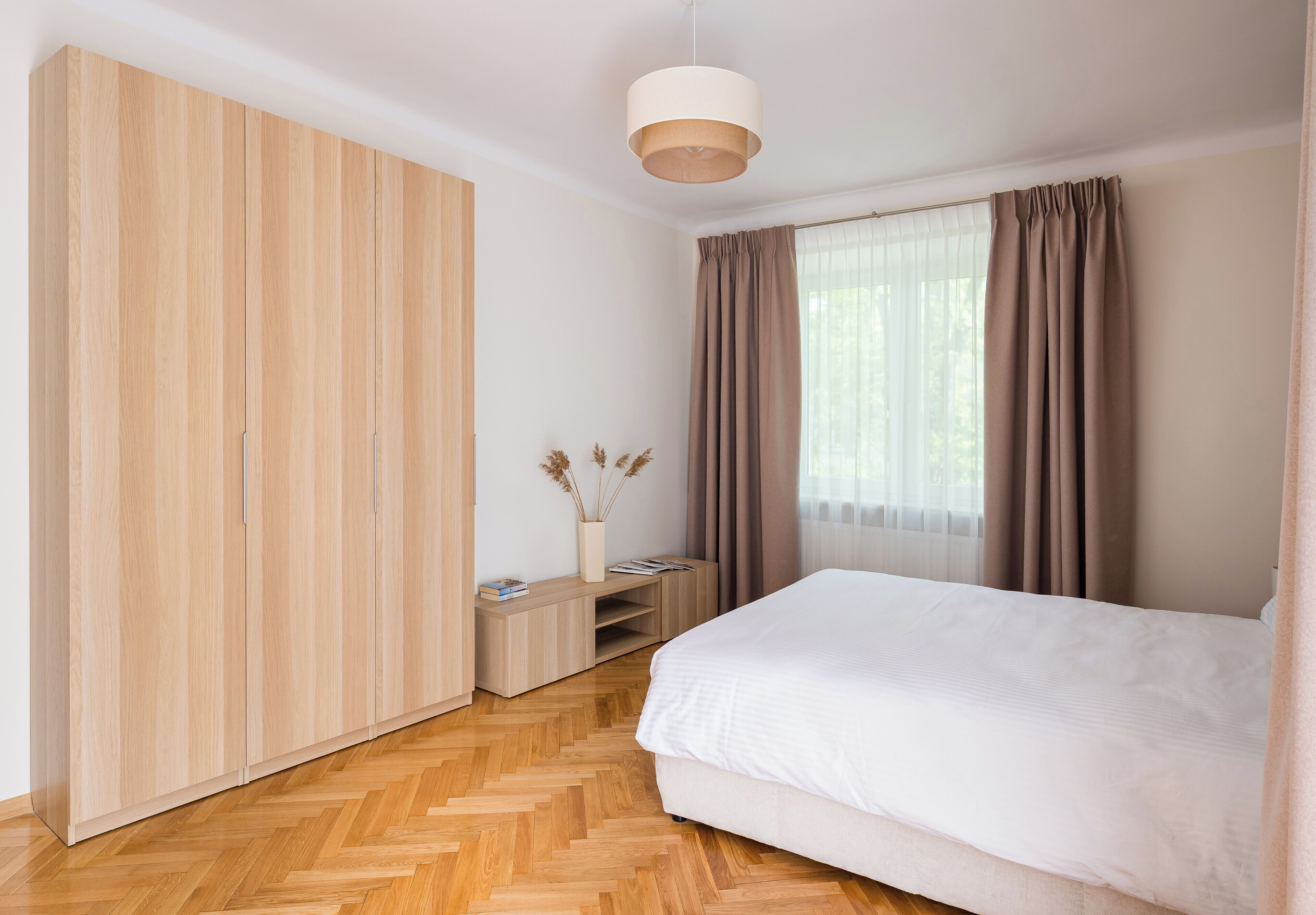 A bedroom with a large wardrobe, a bed, a bedside table with books, and a hanging light fixture.