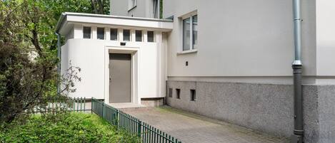 Un edificio moderno con fachada blanca, una pequeña entrada con puerta y un camino pavimentado con valla verde.