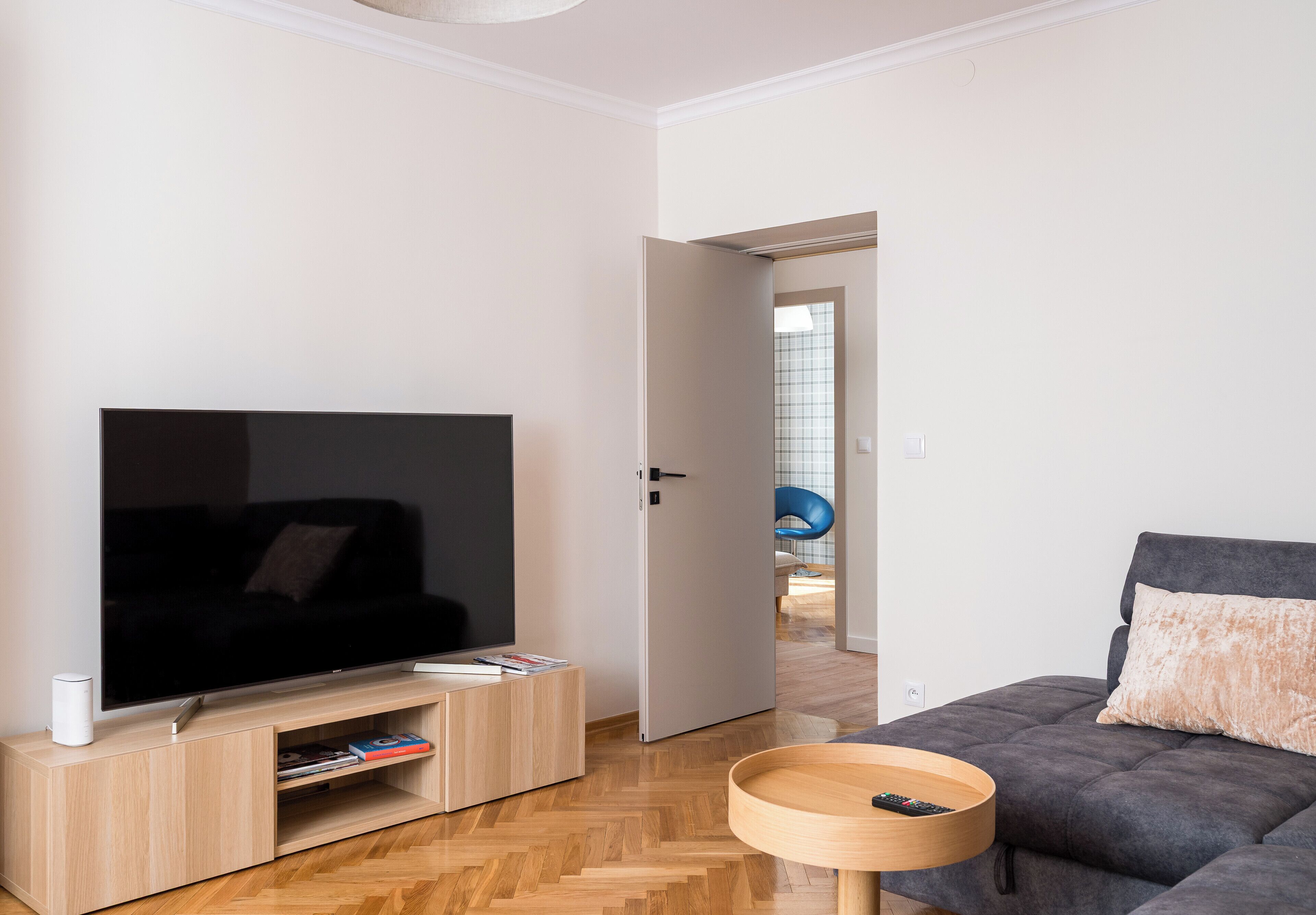 Uma sala de estar moderna com uma TV de tela plana em um suporte de madeira, um sofá cinza com uma almofada e uma pequena mesa de madeira com um controle remoto.