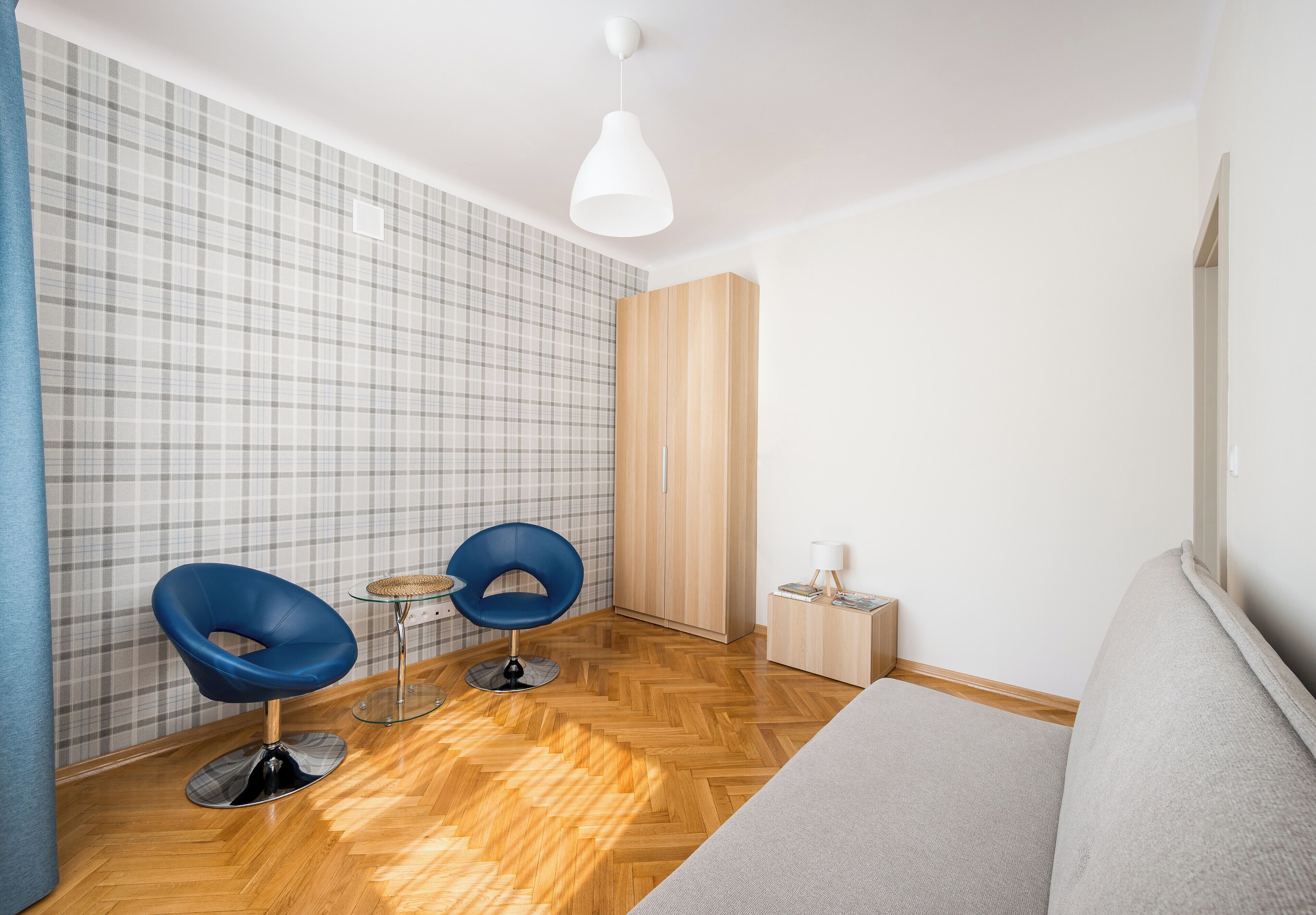 Quarto moderno com piso de madeira em padrão herringbone, duas cadeiras azuis, uma mesa redonda pequena, um guarda-roupa de madeira e um sofá cinza.