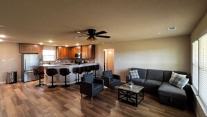 A modern kitchen with wooden cabinets, a central island, and bar stools. There is a living area with a sofa and a coffee table. - Cozy 3BR Home in Poolville / AC + Fast WiFi / Relaxing Escape (Poolville)
