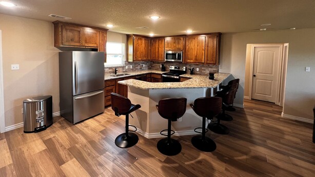 A modern kitchen with wooden cabinets, a stainless steel refrigerator, a microwave, and a granite countertop island with bar stools. - Cozy 3BR Home in Poolville / AC + Fast WiFi / Relaxing Escape (Poolville)