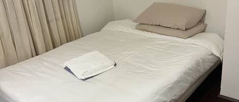 A small, neatly made bed with white bedding and a beige pillow, a wooden floor, and a curtain on the left side.