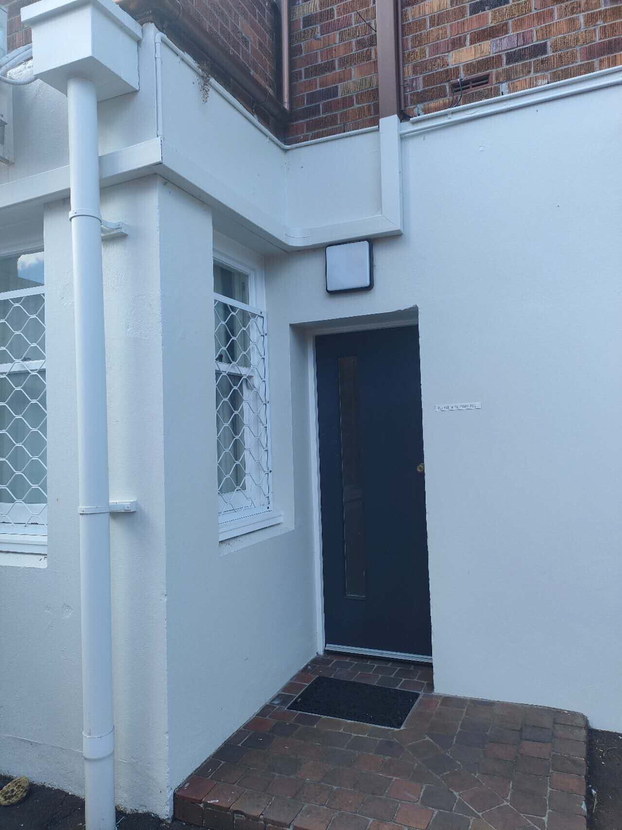 A white building with a dark door, a window with a metal grille, and a small brick-paved area in front.