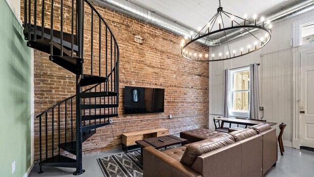Ein modernes Wohnzimmer mit Backsteinwand, einer schwarzen Wendeltreppe, einem Ledersofa, einem Holztisch und einer großen Kronleuchter.
