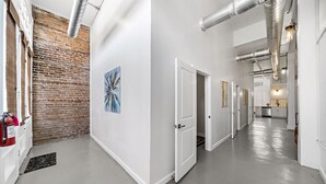 Ein Flur mit weißen Wänden, einer Ziegelwand als Akzent und einem blauen abstrakten Gemälde. Mehrere Türen führen zu Zimmern, und am Ende ist eine Küchenzeile erkennbar.