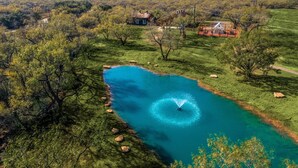 Un extenso cuerpo de agua con una fuente, rodeado de árboles y una casa al fondo.