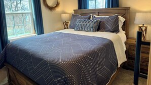 Un dormitorio con una cama grande, dos mesitas de noche, una mesita auxiliar, una ventana con cortinas y una guirnalda en la pared.