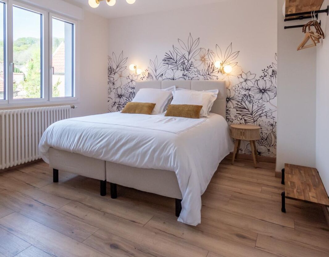 A bedroom with a bed, a nightstand, a wooden bench, and a wall with a floral pattern.
