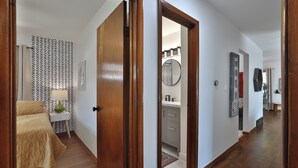 Un couloir avec un plancher en bois, un lit recouvert d’une couvre-lit à motifs, une table de chevet avec une lampe, et un mur orné d’un miroir et d’œuvres d’art.