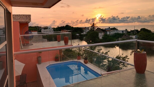 Un balcon avec une piscine, offrant une vue sur un coucher de soleil se reflétant sur une étendue d’eau, avec des bâtiments en arrière-plan.