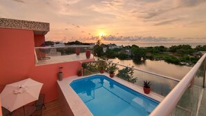 Une piscine sur le toit, entourée d’une balustrade en verre, un espace repas avec une table et des chaises, et une vue sur le coucher de soleil au-dessus d’une étendue d’eau.