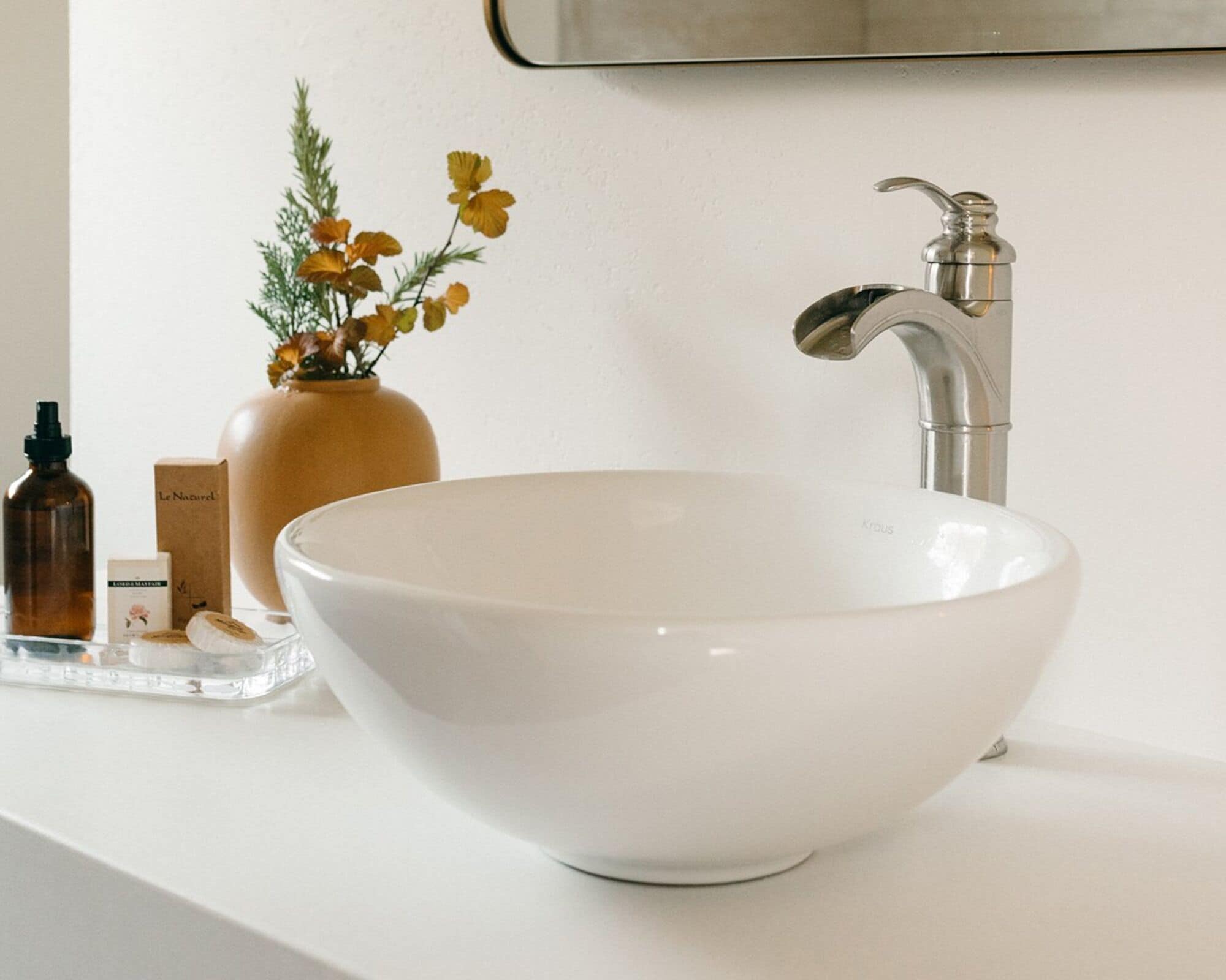 Ein weißes Keramikbecken mit modernem Wasserhahn, einem klaren Glastablett, einer braunen Vase mit gelben Blumen und einigen Pflegeprodukten auf der Arbeitsfläche.