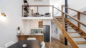 A modern kitchen with a central island, a dining area with a black table, and a staircase leading to an upper level with seating. - Woolstore Style! ~ 2Bed/2Bath/1Car ~ Teneriffe (Teneriffe)