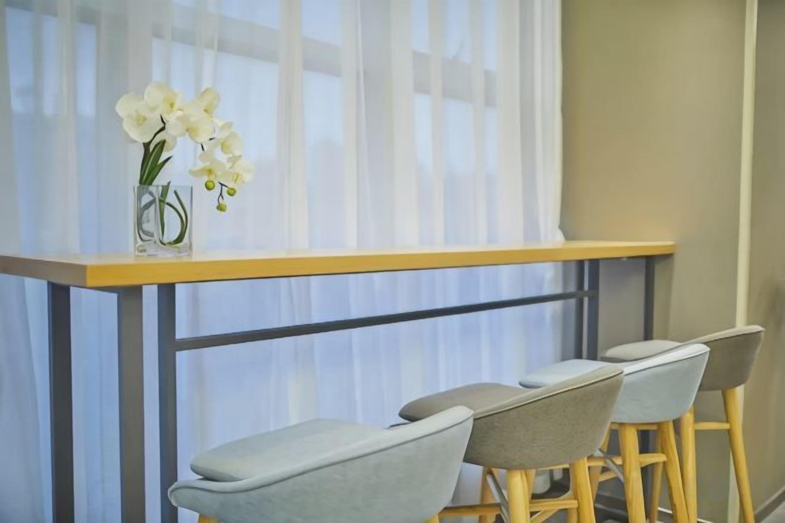 A dining area with a table and chairs, a vase with flowers, and a window with sheer curtains.