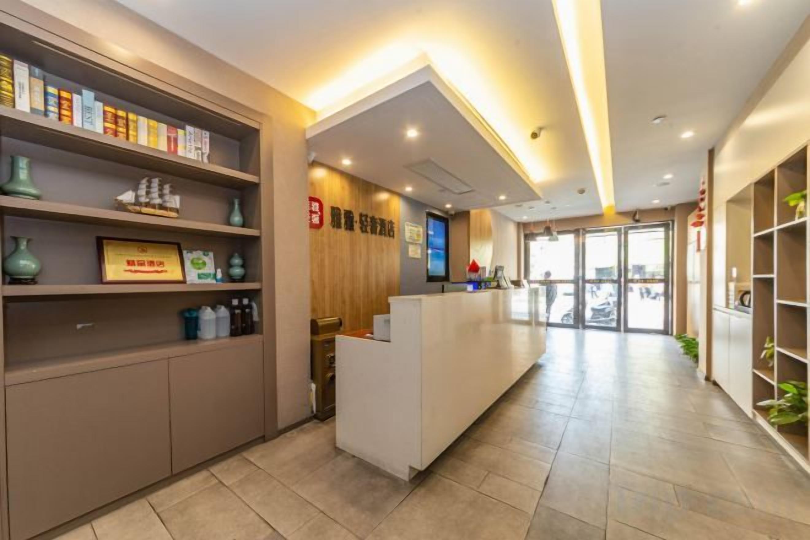 A hotel reception area with a reception desk, shelves with books and decorative items, a wooden wall with signage, and a tiled floor.