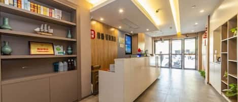 A hotel reception area with a reception desk, shelves with books and decorative items, a wooden wall with signage, and a tiled floor.