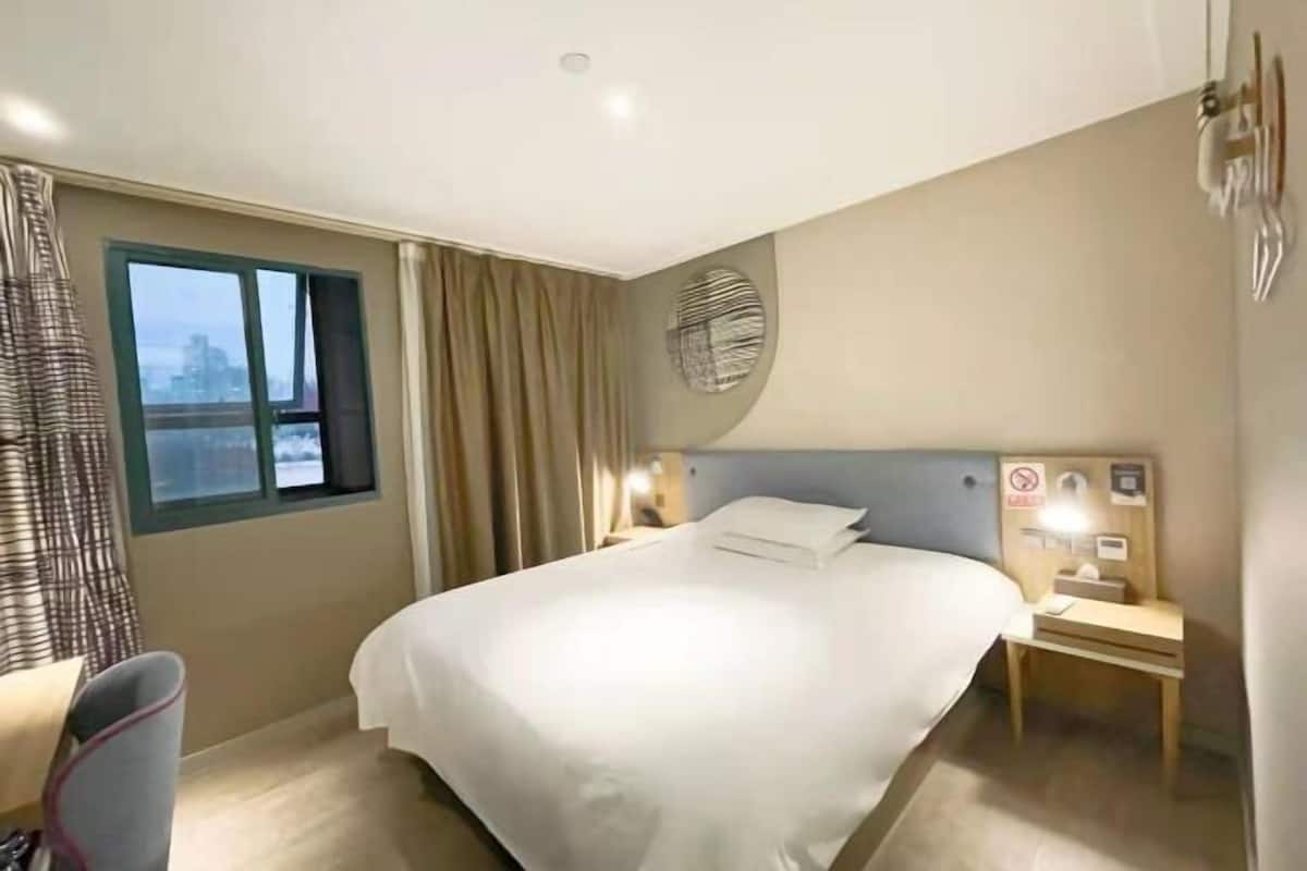 Ein Hotelzimmer mit einem großen Bett, einem Nachttisch, Vorhängen am Fenster und Blick auf die Stadt.