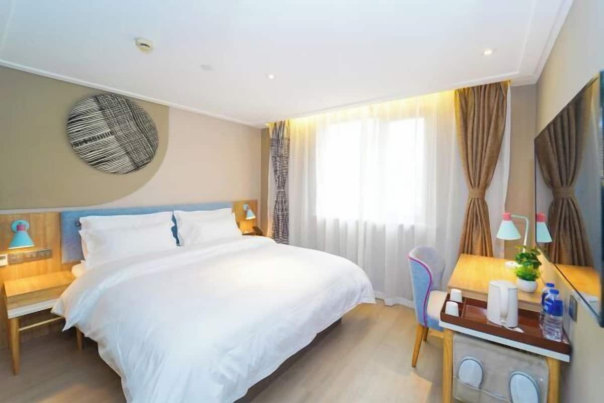 Ein Hotelzimmer mit Bett, Nachttischen, Schreibtisch mit Stuhl, Spiegel und einem Fenster mit Vorhängen.