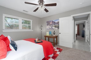 Un dormitorio con cama, ventilador de techo, ventana con persianas, mesita de noche y puerta que da a otra habitación.
