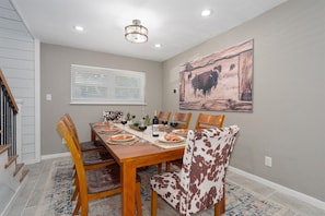 A dining area with a wooden table set for four, surrounded by chairs, a large painting on the wall, and a window with blinds. - Abilene Icon- Sky, Story, and the Western Spirit! (Abilene)