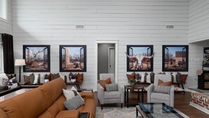 Uma sala de estar ampla com um sofá marrom, poltronas e uma mesa de centro. Há quadros nas paredes e uma porta que dá para outro cômodo.