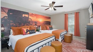 Quarto com duas camas, um ventilador de teto, um mural grande na parede, uma janela com cortinas e uma cômoda.