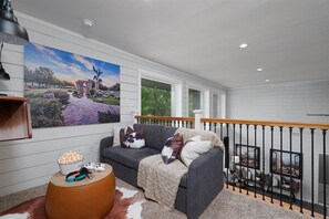 Ein gemütliches Wohnzimmer mit einem grauen Sofa, einem kleinen runden Couchtisch mit Popcorn und einer Fernbedienung, einem großen Gemälde an der Wand und Blick ins Freie durch ein Fenster.