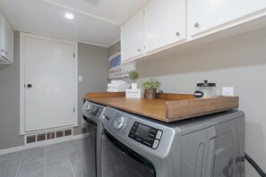 A laundry room with a front-loading washing machine, a dryer, and a wooden shelf with folded towels and a potted plant. - Abilene Icon- Sky, Story, and the Western Spirit! (Abilene)