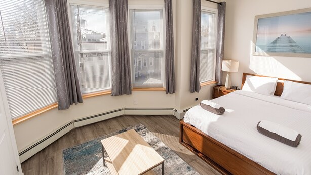 Un dormitorio con una cama grande, una mesita de noche, una lámpara y vistas a la ciudad a través de las ventanas.