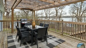Eine überdachte Terrasse im Freien mit Grill, Esstisch und Stühlen, mit Blick auf ein Gewässer.