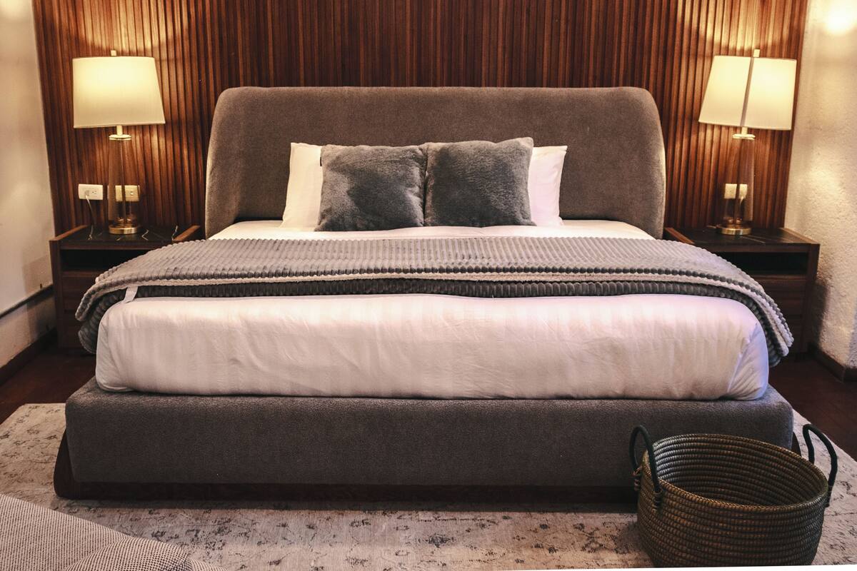 Una cama bien hecha con un edredón gris y blanco, dos lámparas de noche, un cabecero de madera y una cesta tejida en el suelo.