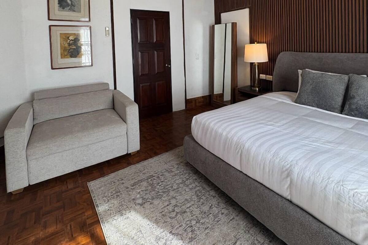 Un dormitorio con cama, sofá, piso de madera, puerta y cuadros en la pared.