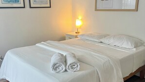 Una cama bien tendida con sábanas blancas y dos toallas enrolladas, una lámpara de noche y cuadros en las paredes.
