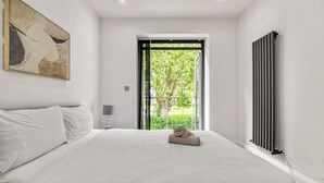 A bedroom with a large bed, a window with a view of greenery, a radiator, and a painting on the wall. - Spacious Modern House 3BR 3BA Balcony Park View (London)