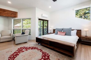 A bedroom with a large bed, a sofa, a chair, a wooden ceiling, an air conditioning unit, and a window with a view of greenery.