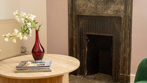 A room with a round wooden table, a red vase with white flowers, and a fireplace with a dark, textured surround. - 1BR, Whitechapel, Brick Lane, Spitalfields (London)