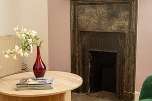 A room with a round wooden table, a red vase with white flowers, and a fireplace with a dark, textured surround.