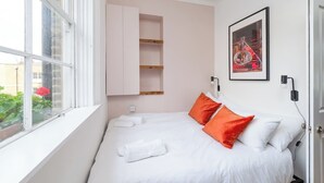 A bedroom with a bed, white bedding, orange pillows, a window, and a framed picture on the wall. - Stunning luxury flat in Prime Central London (London)