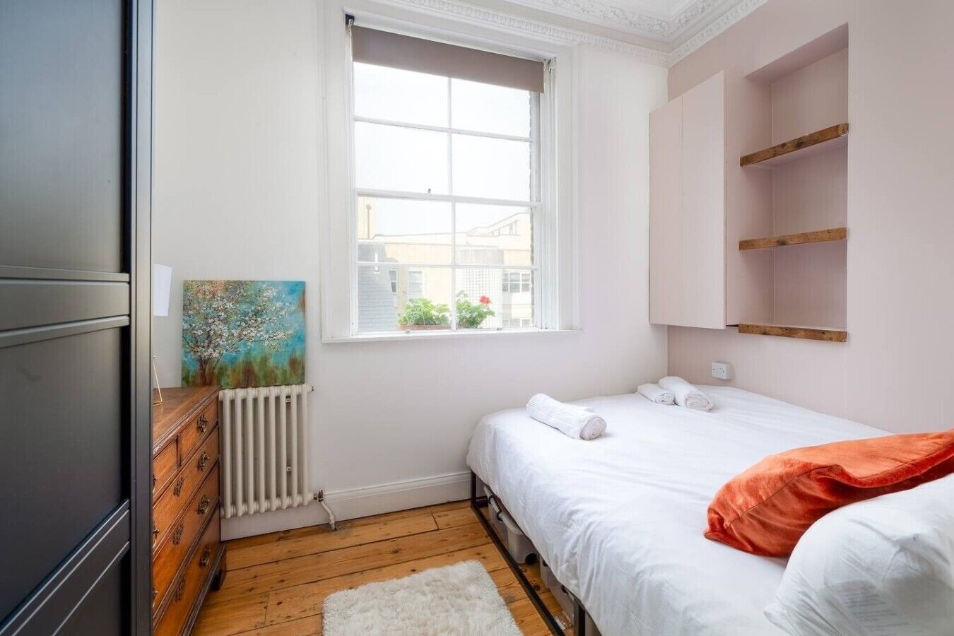 A bedroom with a bed, a radiator, a window, a painting, and a wardrobe.