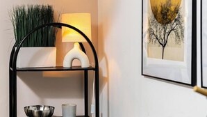 Habitación de esquina con una mesa blanca, una silla negra, un estante metálico con un cuenco y una vela, una lámpara y un cuadro en la pared.