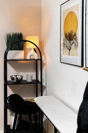 Ein Eckzimmer mit einem weißen Tisch, einem schwarzen Stuhl, einem Metallregal mit Schüssel und Kerze, einer Lampe und einem gerahmten Bild an der Wand.