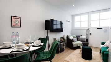 A modern living room with a dining area, green chairs, a flat-screen TV, and a large window.